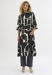 Donna con capelli ricci che indossa un lungo cappotto astratto nero e beige, jeans scuri e sandali bianchi aperti, in piedi davanti a uno sfondo bianco.