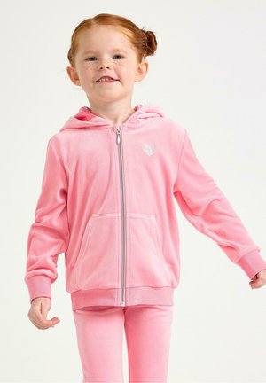 Ung pige med rødt hår i fletninger iført en lyserød hættetrøje med lynlås og et lille hjerteemblem samt matchende lyserøde bukser, smilende.