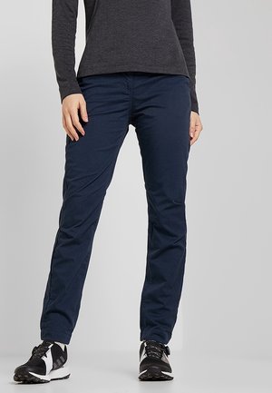 Persona que lleva pantalones azul oscuro, zapatillas blancas y negras y una camiseta de manga larga oscura, de pie frente a un fondo liso.