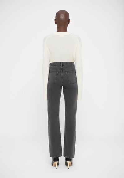 Persona con cabeza rapada que lleva un suéter de punto color crema, jeans de cintura alta gris oscuro y zapatos de tacón negros de pie, de espaldas.