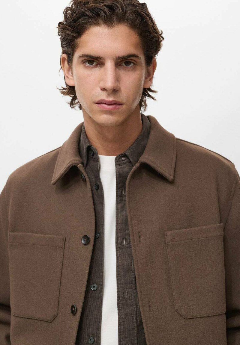 Giovane uomo con capelli corti e mossi che indossa una giacca marrone sopra una camicia scura con bottoni e una maglietta bianca, rivolto in avanti con un'espressione neutra.