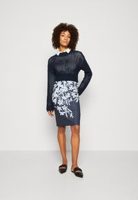 Un pull cropped en tricot bleu marine avec un tissage ouvert, superposé à une jupe foncée ajustée ornée de motifs floraux blancs. Des chaussures plates noires complètent l'ensemble.