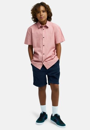 Kurzärmeliges, hellrosa Hemd mit braunen Knöpfen, kombiniert mit marineblauen Shorts und schwarzen Sneakern mit türkisfarbenen Akzenten und weißen Socken.