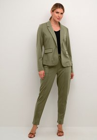 Alyvuogių žalios spalvos pritaikytas kostiumas su aptempta švarku, turinčiu dvi priekinės kišenes, derinamas su siaurėjančiomis kelnėmis. Lygi, lengva medžiaga.