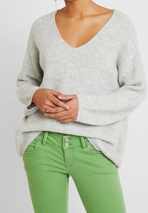 Maglione lavorato a maglia grigio chiaro con scollo a V profondo, maniche ampie e vestibilità rilassata, abbinato a jeans verdi attillati con chiusura a bottone.
