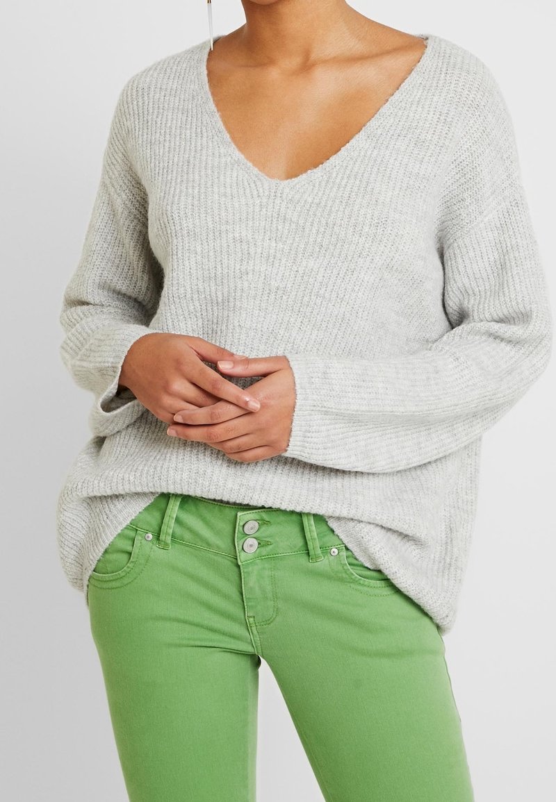 Helles graues Strickoberteil mit tiefem V-Ausschnitt, weiten Ärmeln und lässiger Passform, kombiniert mit figurbetonten grünen Jeans mit Knopfverschluss.