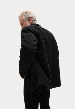 Schwarze Cordjacke mit vertikaler Rippstruktur, die einen lockeren Schnitt, seitliche Taschen und geknöpfte Manschetten aufweist. Hinten leicht geöffnet getragen.