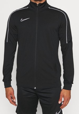 Mann trägt eine schwarze Nike-Jacke mit Reißverschluss und weißen Paspeln an den Ärmeln sowie passende schwarze Shorts mit Nike-Logo am linken Bein.