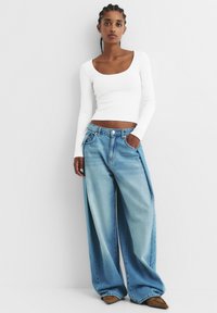 Un haut long à manches blanches en maille côtelée associé à un jean en denim large couleur bleu clair. Le jean possède une fermeture à bouton et des poches avant. Chaussures brunes à bout pointu.