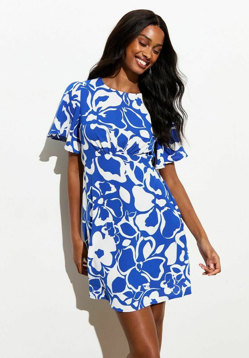 Mujer sonriente con largo cabello negro que lleva un vestido de manga corta azul y blanco con flores, de pie frente a un fondo blanco liso.