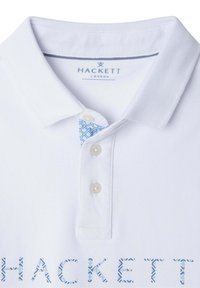 Biela polokošeľa s textúrovanou látkou, klasickým golierom, tromi gombíkmi a modrým vzorovaným akcentom vo vnútri goliera. Logo "HACKETT" v modrej farbe na spodnej časti.
