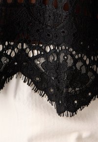 Tissu en dentelle noire avec des motifs floraux et géométriques complexes, drapé sur un fond blanc texturé.
