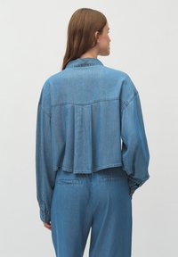 Svetlo modra denim jakna z udobnim fitom, spuščena ramena in detajlom z gubami na hrbtu. Dolgi rokavi z gumbi na manšetah in ovratnik.