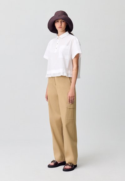 Camisa polo blanca recortada con un dobladillo rizado, combinada con pantalones de pierna ancha color marrón claro que tienen un bolsillo lateral. Un sombrero tipo cubo marrón y sandalias negras completan el look.