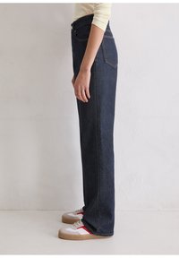 Hochtaillierte, dunkelblaue Jeans mit geradem Bein. Verfügt über klassische Taschen und kontrastierende Nähte. Getragen mit weißen Sneakern.