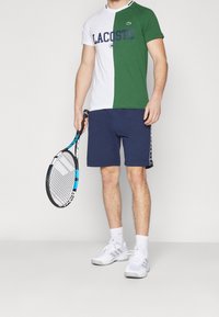 Man wearing half-green, half-white Lacoste T-shirt, navy shorts, white sneakers, holding a black and blue tennis racket against a plain background.