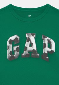 Zelena bombažna majica s kratkimi rokavi in okrolim izrezom, na kateri so velike črke "GAP" oblikovane z vzorcem nogometne žoge.