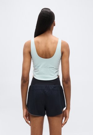 Naisel pikad tumedad juuksed, kandes helesinist varrukateta lühikest toppi ja musti spordipükse, seisab näoga eemale hallil taustal.