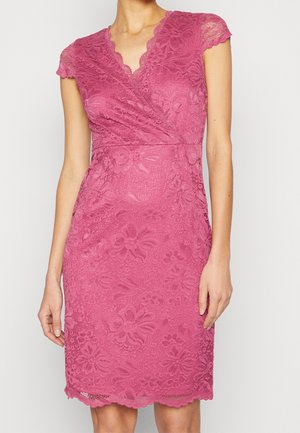 Mujer con un vestido rosa de encaje hasta la rodilla, con mangas cortas y escote en V, de pie frente a un fondo liso y claro.