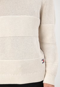 Suéter de punto, beige claro con patrones texturizados y puños acanalados. Presenta una etiqueta de marca. Diseñado con detalles de rayas horizontales.