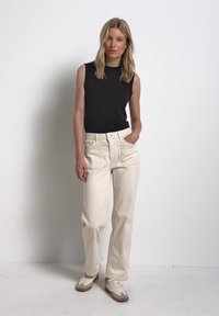 Frau mit blondem Haar, die ein ärmelloses schwarzes Top, beige hoch taillierte Hose und weiße Sneakers trägt und mit einer Hand in der Tasche steht.