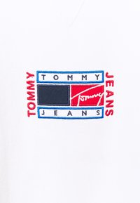 Tessuto bianco con scollo a V caratterizzato da un logo ricamato. Il design include elementi rossi, blu e neri con il testo "TOMMY JEANS".