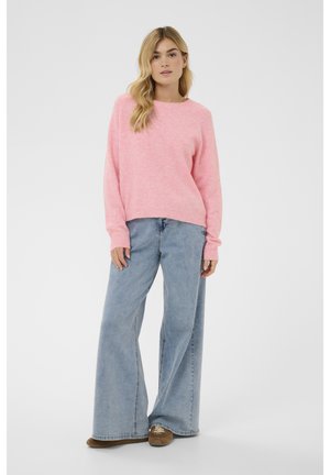 Mladá žena stojí, oblečená v měkkém růžovém svetru, širokých světle modrých džínách a hnědých teniskách, na jednobarevném bílém pozadí.
