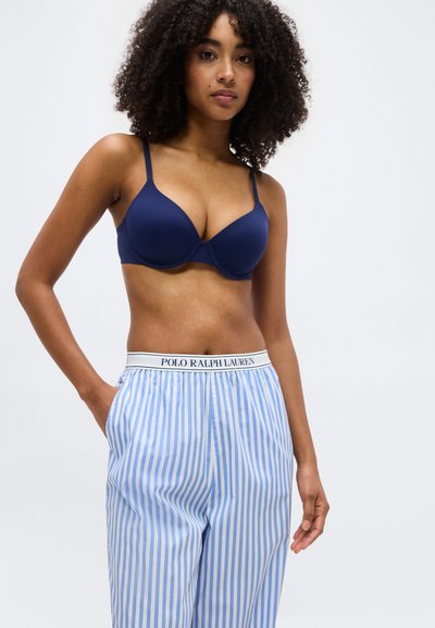 Γυναίκα με σγουρά μαλλιά που φοράει μπλε σουτιέν και μπλε-άσπρο ριγέ πυτζάμα με ζώνη μέσης "Polo Ralph Lauren", με το χέρι στην τσέπη, όρθια.