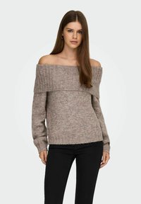 Pull décolleté en beige clair, texture côtelée, avec une coupe ample, des manches longues et un ourlet court. Associé à un pantalon noir.