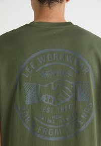 Olíva zöld pamut póló köralakú grafikával, amelyen kézfogás és a "LEE WORKWEAR", "EST 1889" és a "S LEGENDÁKBÓL SZÜLETETT" szöveg szerepel.