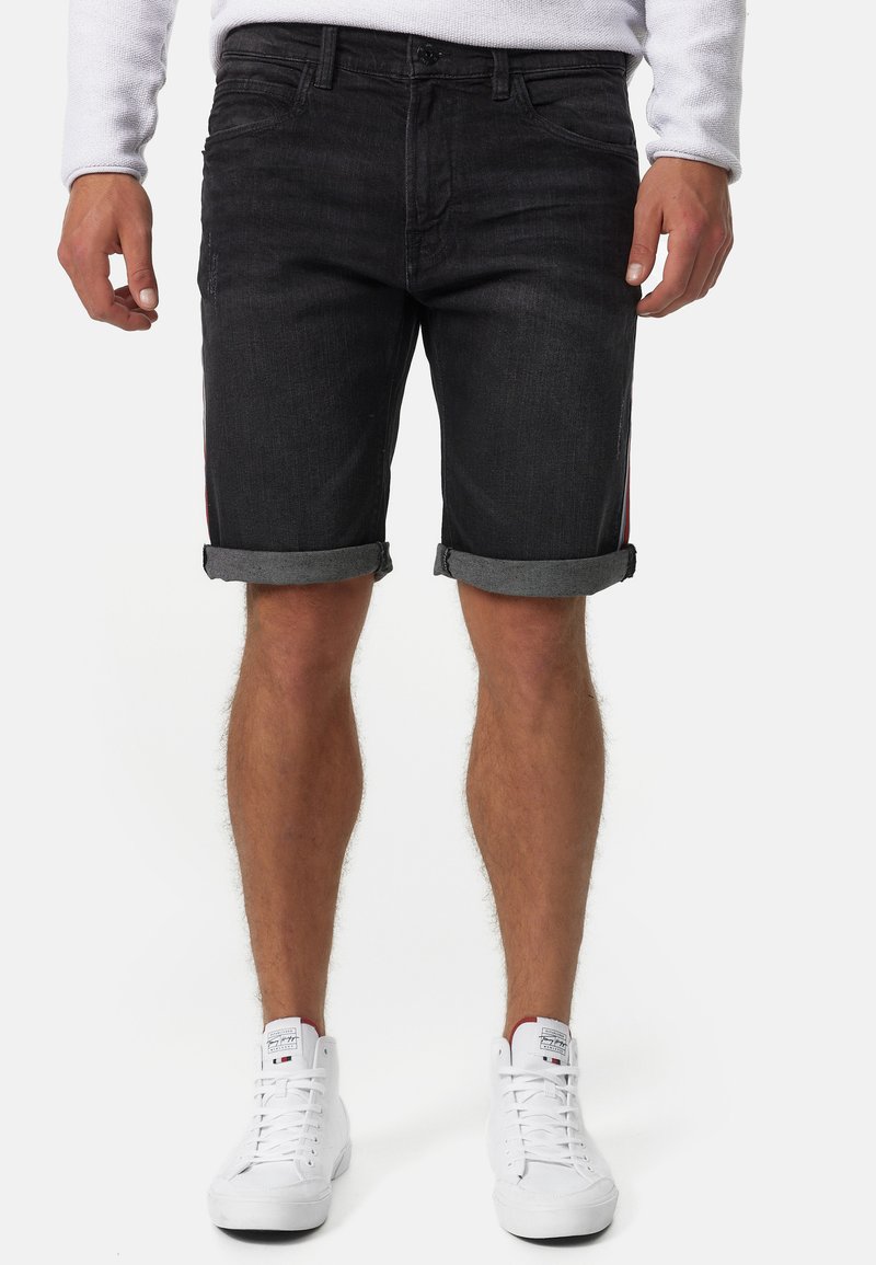 Shorts en denim noir avec des ourlets repliés, présentant un design à cinq poches et des accents rouges discrets sur les côtés. Assortis à des baskets blanches.