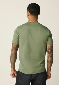 Bavlnené tričko olivovo zelenej farby s okrúhlym výstrihom, krátkymi rukávmi, hladkou textúrou a mierne priliehavým strihom; nosené s tmavými rifľami.