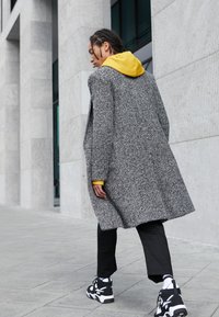 Osoba s copánky chodí venku a má na sobě šedý texturovaný kabát přes žlutou mikinu s kapucí, černé kalhoty a černobílé tenisky.