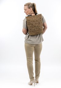 Mujer con pantalones beige, top topo y tacones altos, llevando una mochila marrón con patrón geométrico y la etiqueta "SURI FREY" en la espalda.