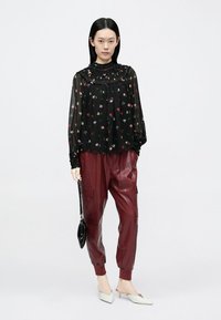 Blusa nera trasparente con motivi floreali, abbinata a pantaloni cargo in pelle bordeaux. Modella con una piccola borsa nera e tacchi bianchi appuntiti.