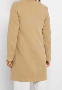 Abrigo de mezcla de lana beige con un corte ajustado, que presenta mangas largas, un cuello alto y un acabado suave, mostrado desde la parte posterior.