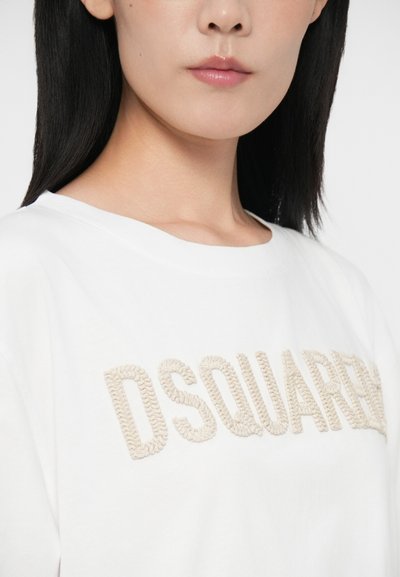 Mladá žena v bielom mikinovom svetri s béžovou textúrovanou nášivkou "DSQUARED2", ukazujúca svoj krk a čiastočne tvár s rovnými čiernymi vlasmi.