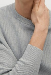 Jasnoszary prążkowany sweter z okrągłym dekoltem i fakturowaną tkaniną. Widoczna dłoń oparta na szyi, ukazująca gładką skórę.