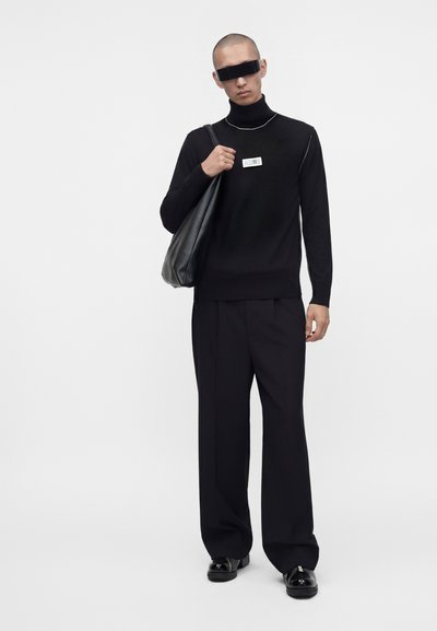 Homme en col roulé noir, pantalon large noir et chaussures noires, portant un grand sac en cuir noir sur l’épaule devant un fond uni.