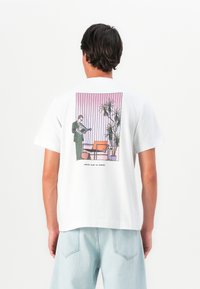 Homem vestindo uma camiseta branca com a imagem de uma pessoa em traje a ler um jornal ao lado de uma cadeira e de uma planta alta, contra um fundo listrado em rosa.
