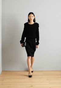 Pull en tricot noir avec un col large, associé à une jupe crayon noire. Le modèle tient un petit sac à main noir et porte des talons noirs.