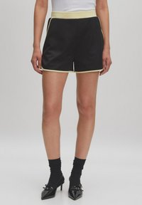 Shorts en satin noir avec une large ceinture jaune et des bordures jaunes sur les côtés. Texture lisse, coupe ajustée et longueur mi-cuisse.