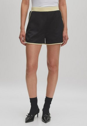 Zwarte satijnen shorts met een brede gele tailleband en gele piping langs de zijkanten. Gladde textuur, op maat gemaakt pasvorm en lengte tot halverwege de dijen.