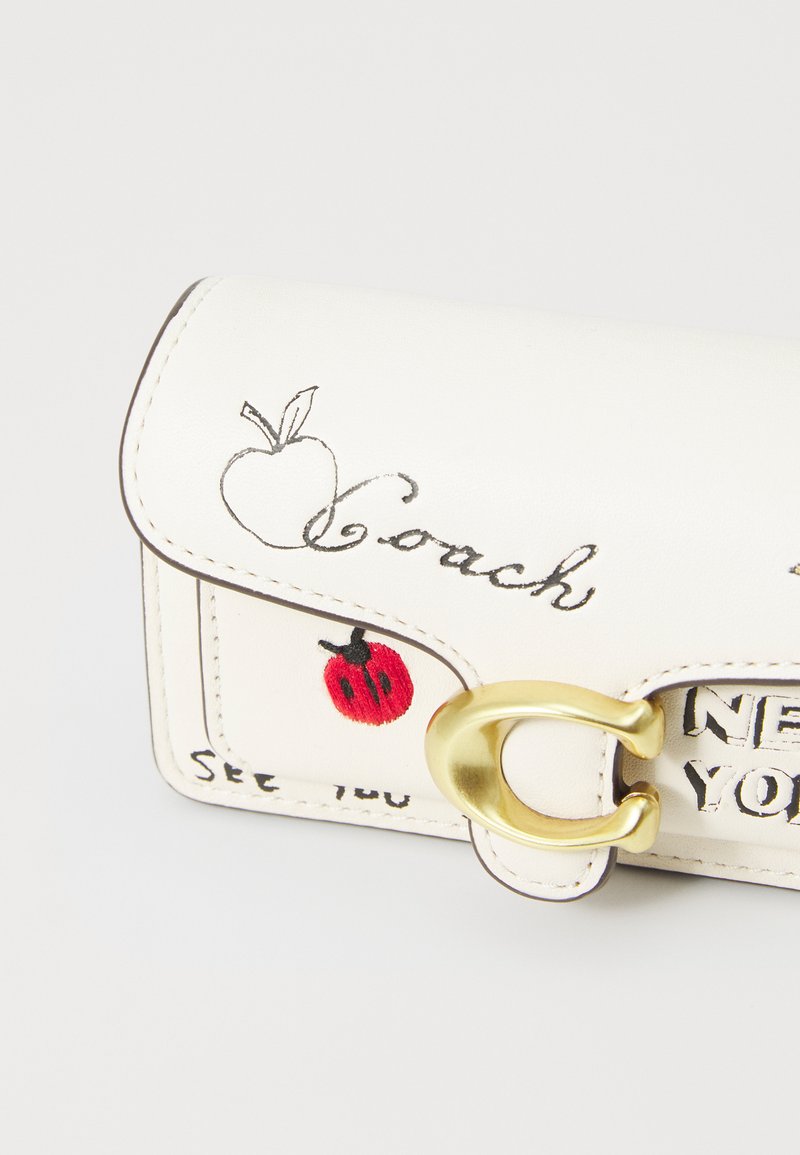 Sac en cuir blanc avec des accessoires en or, présentant des broderies de pomme et de coccinelle, ainsi que le logo "Coach". Texture lisse et forme rectangulaire compacte.