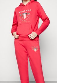 Ensemble sweat à capuche rouge et pantalon de jogging, avec un logo sur la poitrine et la jambe. Matière en coton au toucher lisse. Capuche avec cordon de serrage et poche avant.