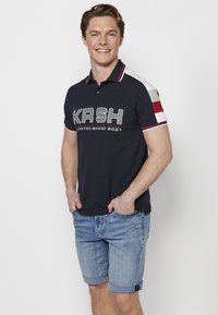 Tengerészkék póló fehér és szürke csíkos ujjal, kiemelkedő "KRSH" logóval, világoskék farmer rövidnadrággal párosítva.
