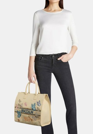 Donna con top bianco a maniche lunghe e jeans neri che tiene una borsa beige con disegni di farfalle blu e rosa e logo "BRACCIALINI".