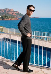 Homme en pull gris, pantalon noir et lunettes de soleil, debout les mains sur les hanches près d'une piscine surplombant l'océan et des collines rocheuses au soleil.