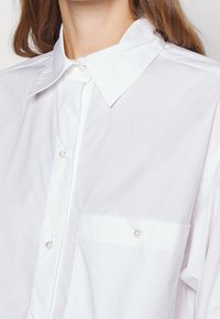 Camicia bianca realizzata in tessuto leggero, con colletto a punta, chiusura con bottoni e una singola tasca sul petto con un dettaglio di bottone perla.