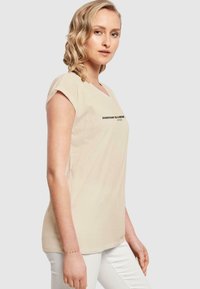 Beige bomull t-skjorte med korte ermer, med V-hals og svart tekst "HVER DAG ER EN NY..." vist på forsiden. Enkelt design.